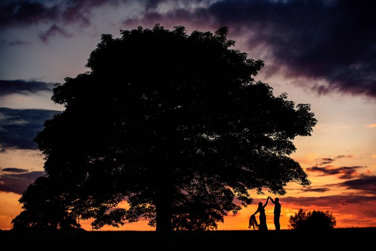 beautiful orange & purple sunset photo as bride twirls with her dress in one hand and next to a large tree at Heaton House Farm