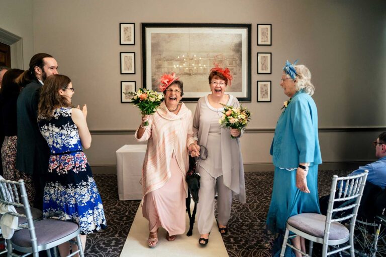 two mature ladies wearing pink fascinators laughing as they have got married and about to walk back down the aisle with their dog, as guests clap.