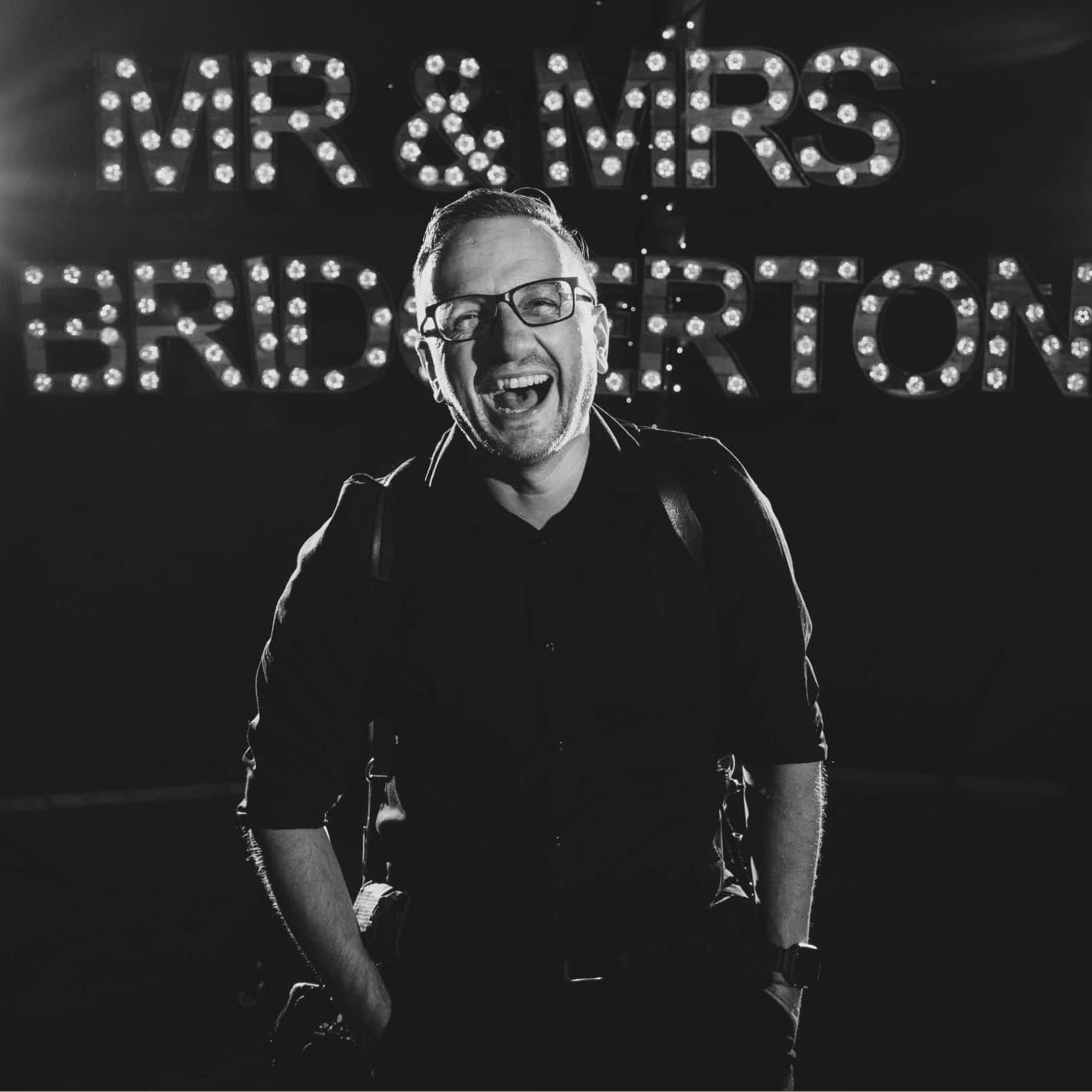 Nick Georgiou, wedding photographer, laughing in front of an illuminated Mr and Mrs light up sign
