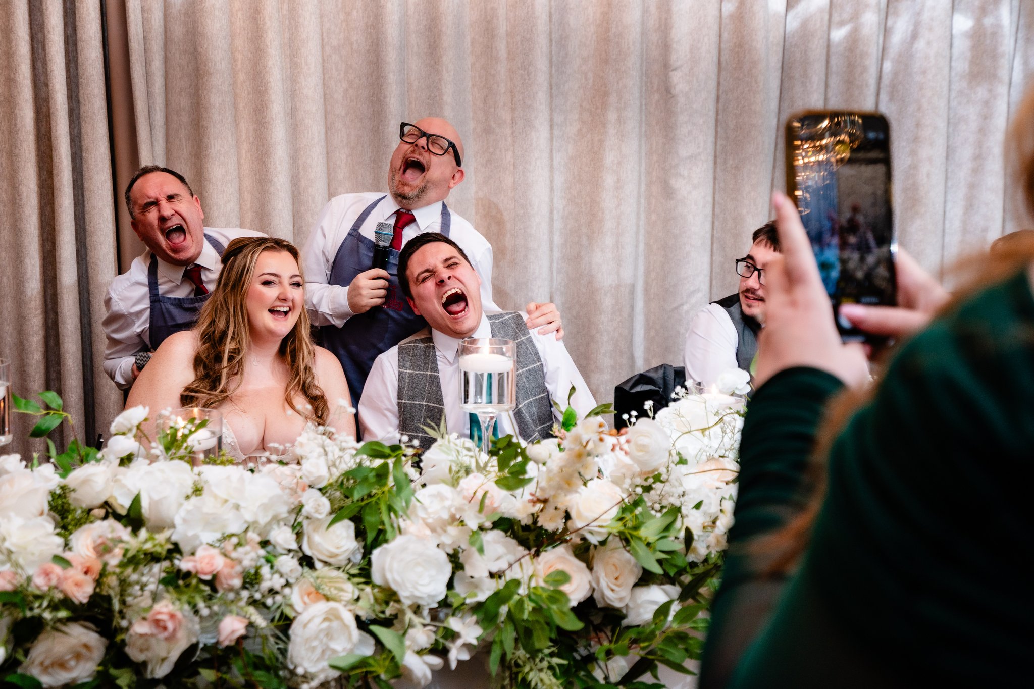 Bride and groom laughing in surprise as a guest films the Singing Waiters on her phone at Merrydale Manor