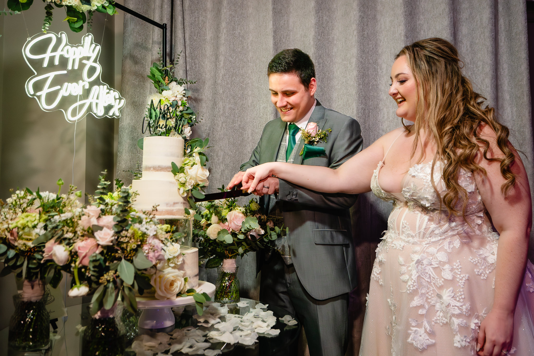 Bride and groom cutting their wedding cake beside a Happily Ever After neon sign at Merrydale Manor