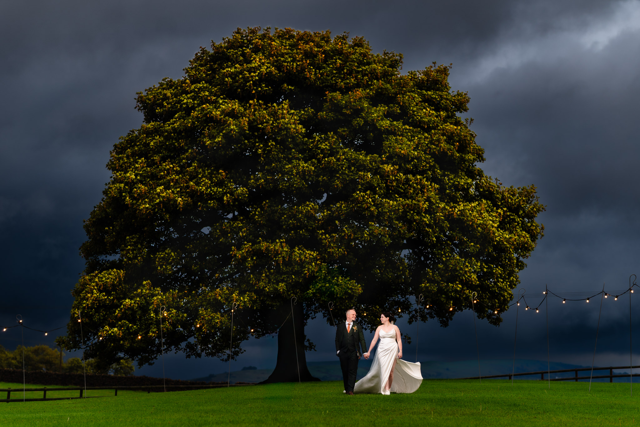 Bride and groom walking hand in hand under the huge oak tree at dusk with a stormy sky above, Heaton House Farm wedding photographer