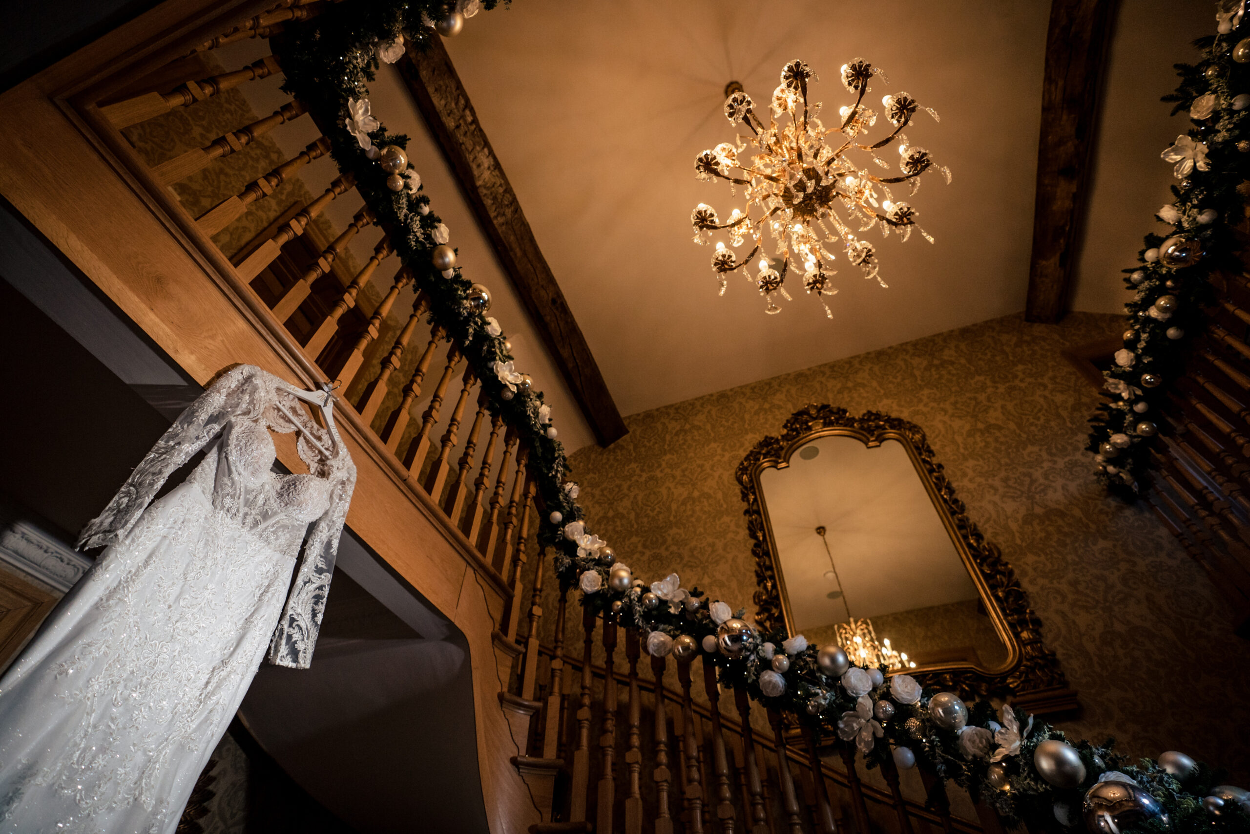 Abbie’s lace wedding dress hanging from a garland wrapped staircase beneath a chandelier, Merrydale Manor wedding photographer