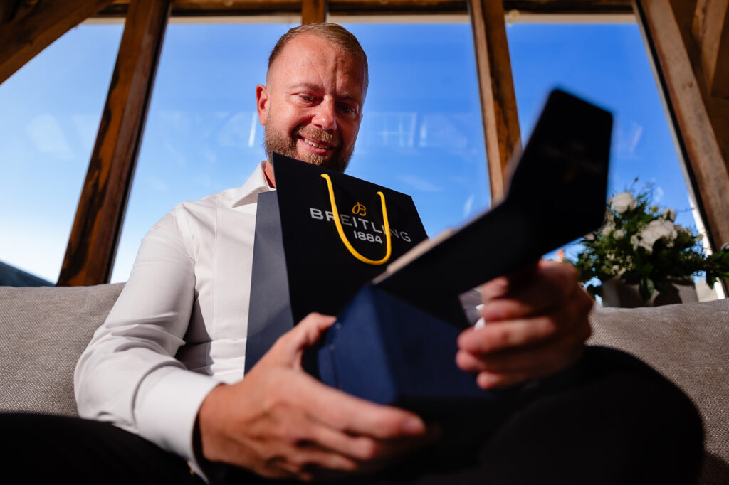 Glynn opening a Breitling gift box on a sofa under oak beams on the wedding morning, Swancar Farm wedding photographer