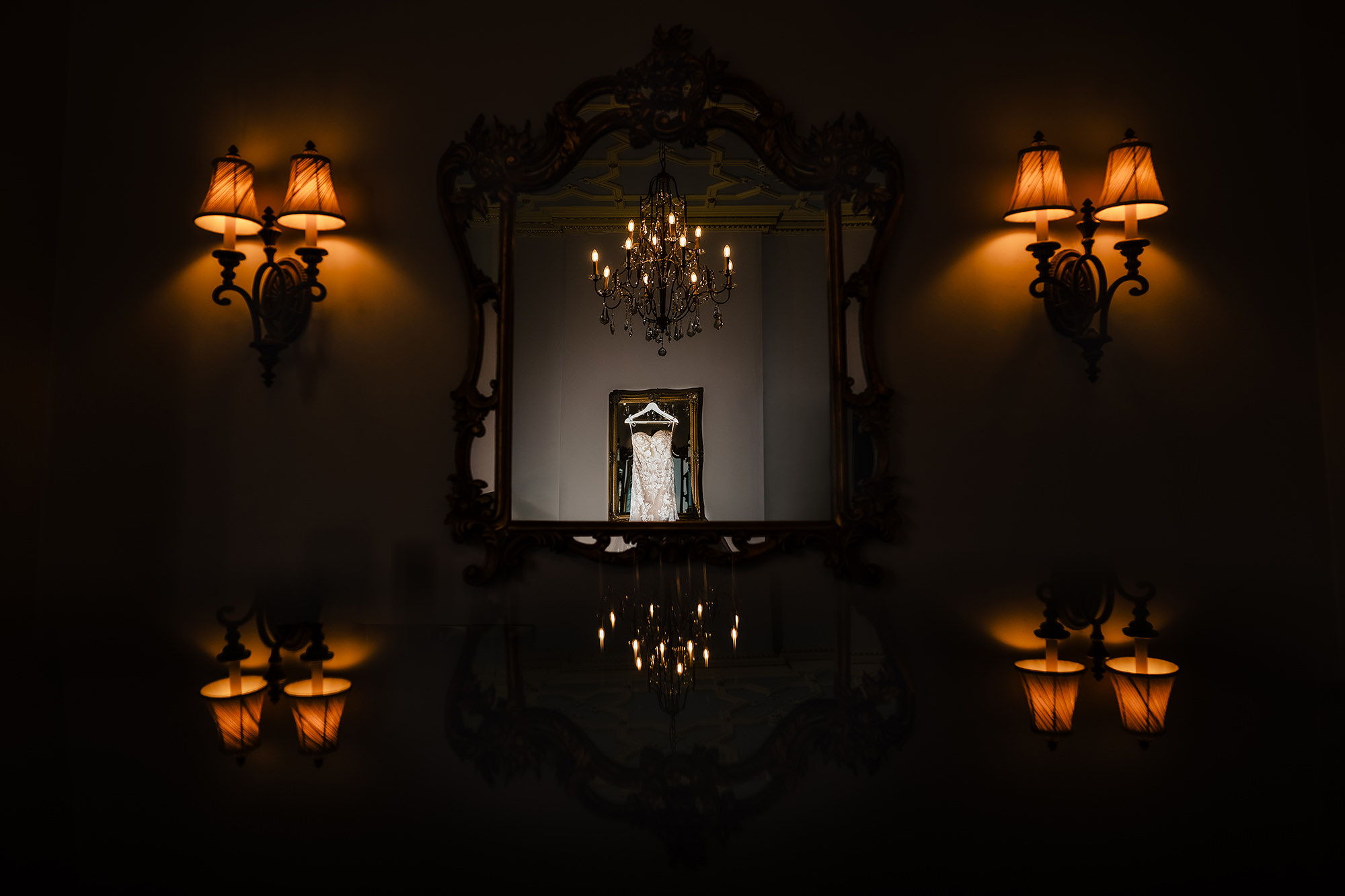 Wedding dress hanging and reflected in an ornate gilt mirror between two lit wall sconces in the bridal suite at Crewe Hall