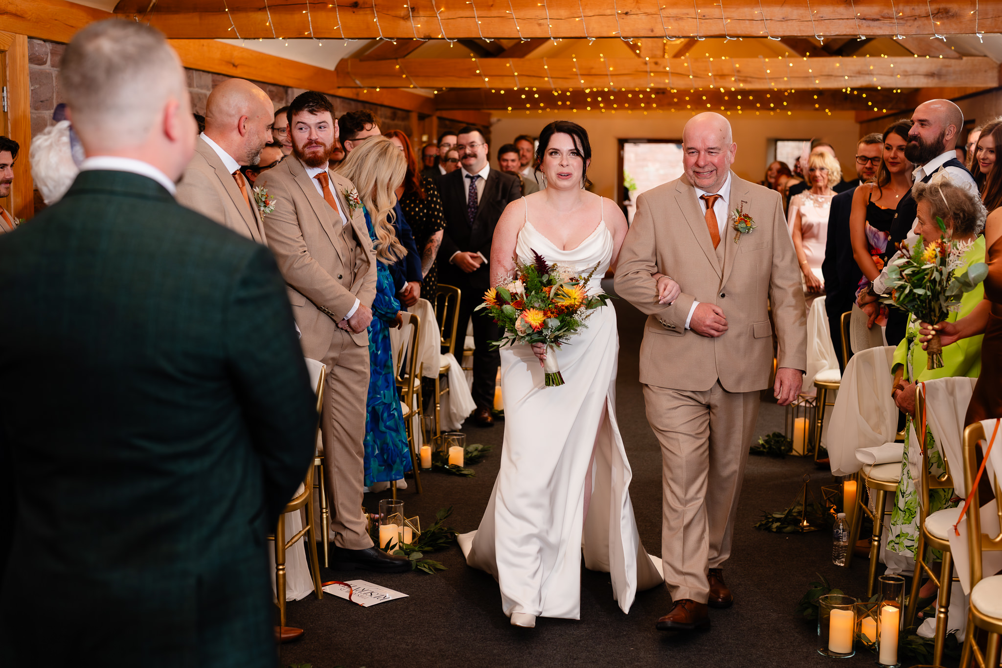 Bride walking down the aisle on her dad's arm holding an orange and yellow autumn bouquet, Heaton House Farm ceremony barn