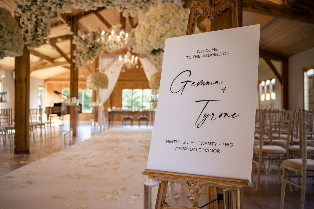 Welcome sign for Gemma and Tyrone at the end of a petal aisle in the Merrydale Manor ceremony room, dressed with white roses and hanging flowers