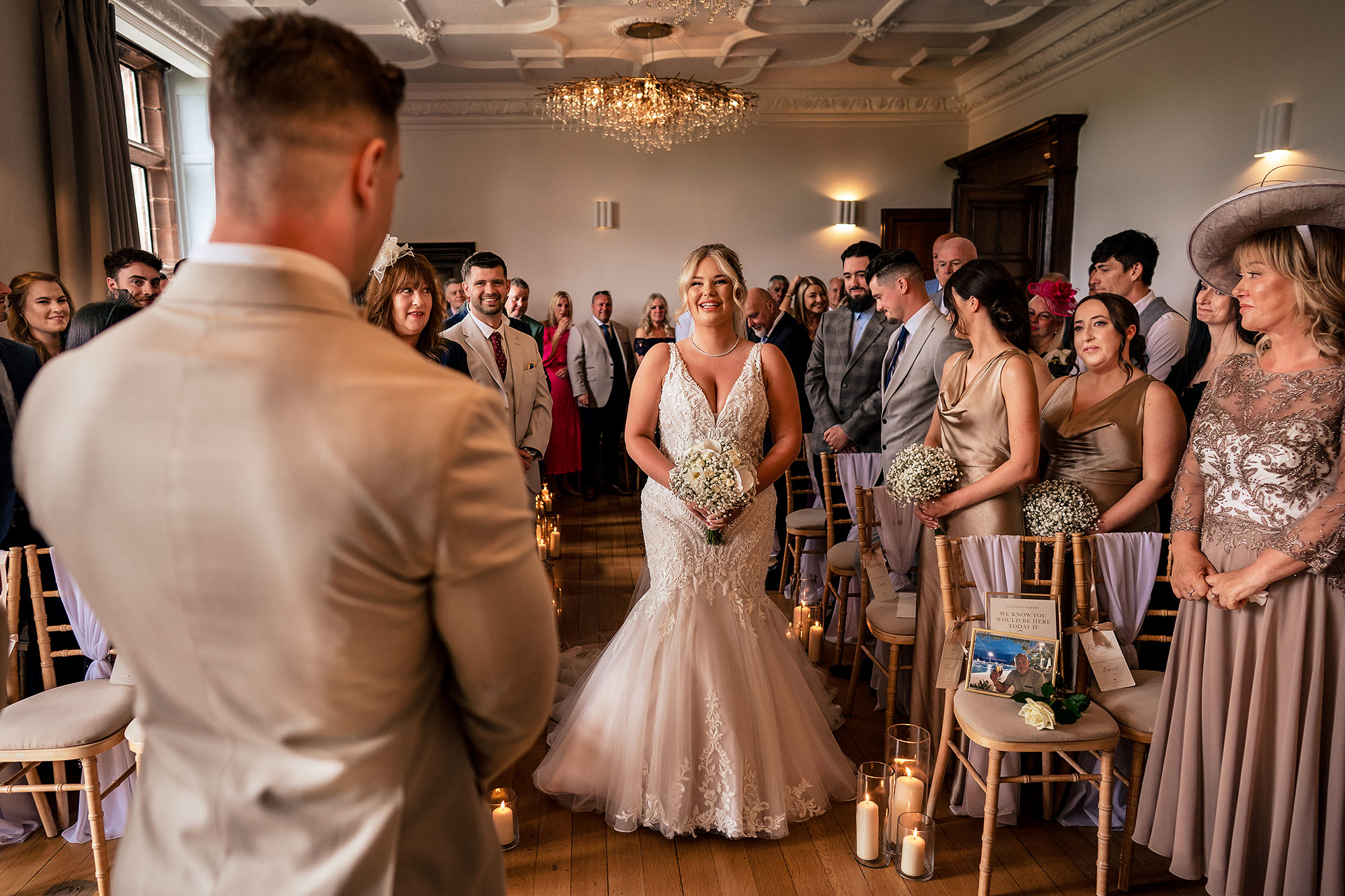 Jess walking into the ceremony room at Salt House, Staffordshire, smiling at Cy as her wedding party watches on