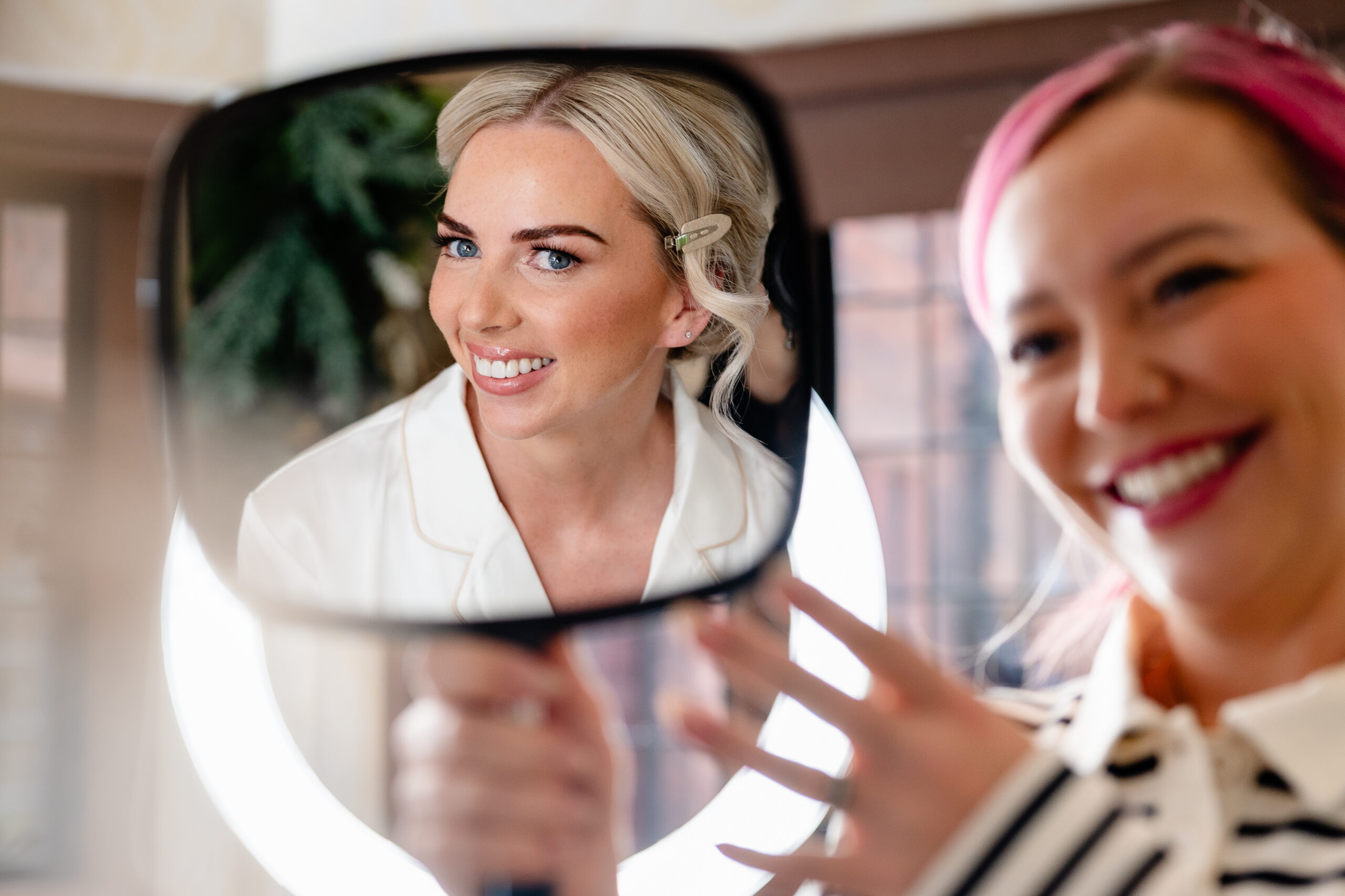Bride smiling at herself in a hand mirror held up by her hair stylist, Colshaw Hall wedding photographer
