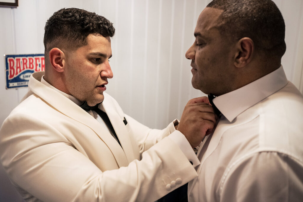 Tyrone in his white tuxedo jacket fastening a bow tie for a guest in the groom’s prep room at Merrydale Manor