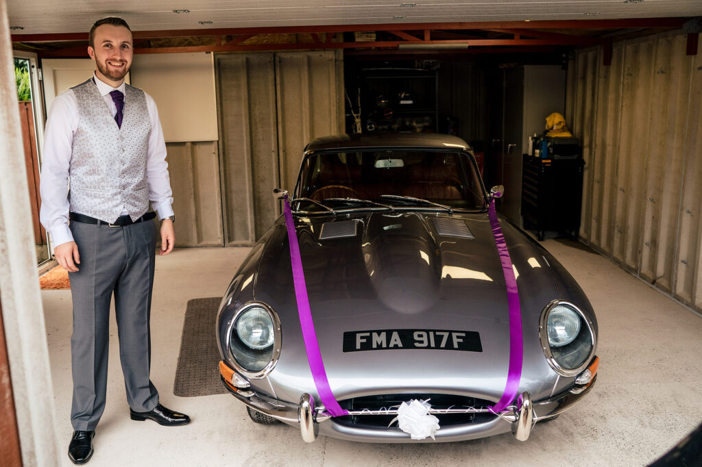 Groom in his waistcoat standing beside the restored silver E-type Jaguar dressed in purple ribbon, Merrydale Manor wedding photographer