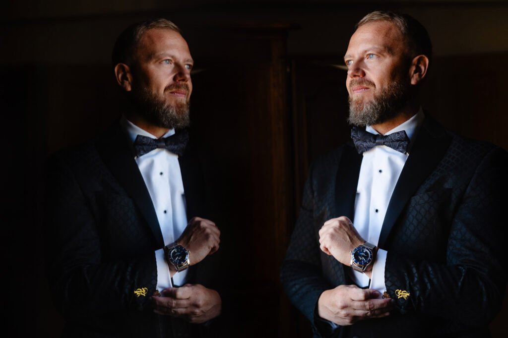 Glynn in a black patterned dinner jacket and bow tie beside his reflection in a mirror, dramatic low light, Nottingham wedding photographer