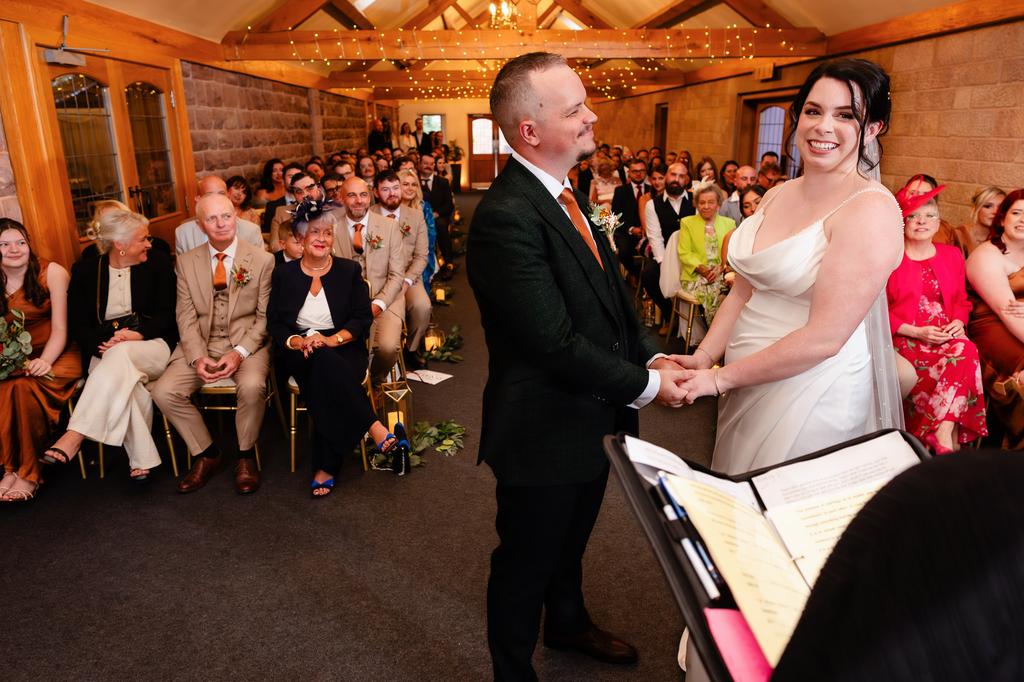 Bride and groom holding hands and laughing during their vows in front of their guests, Heaton House Farm Cheshire wedding photographer