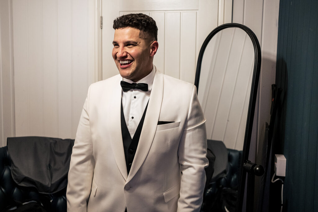 Tyrone laughing in a white tuxedo jacket and black bow tie, ready for his Merrydale Manor wedding, Cheshire wedding photographer