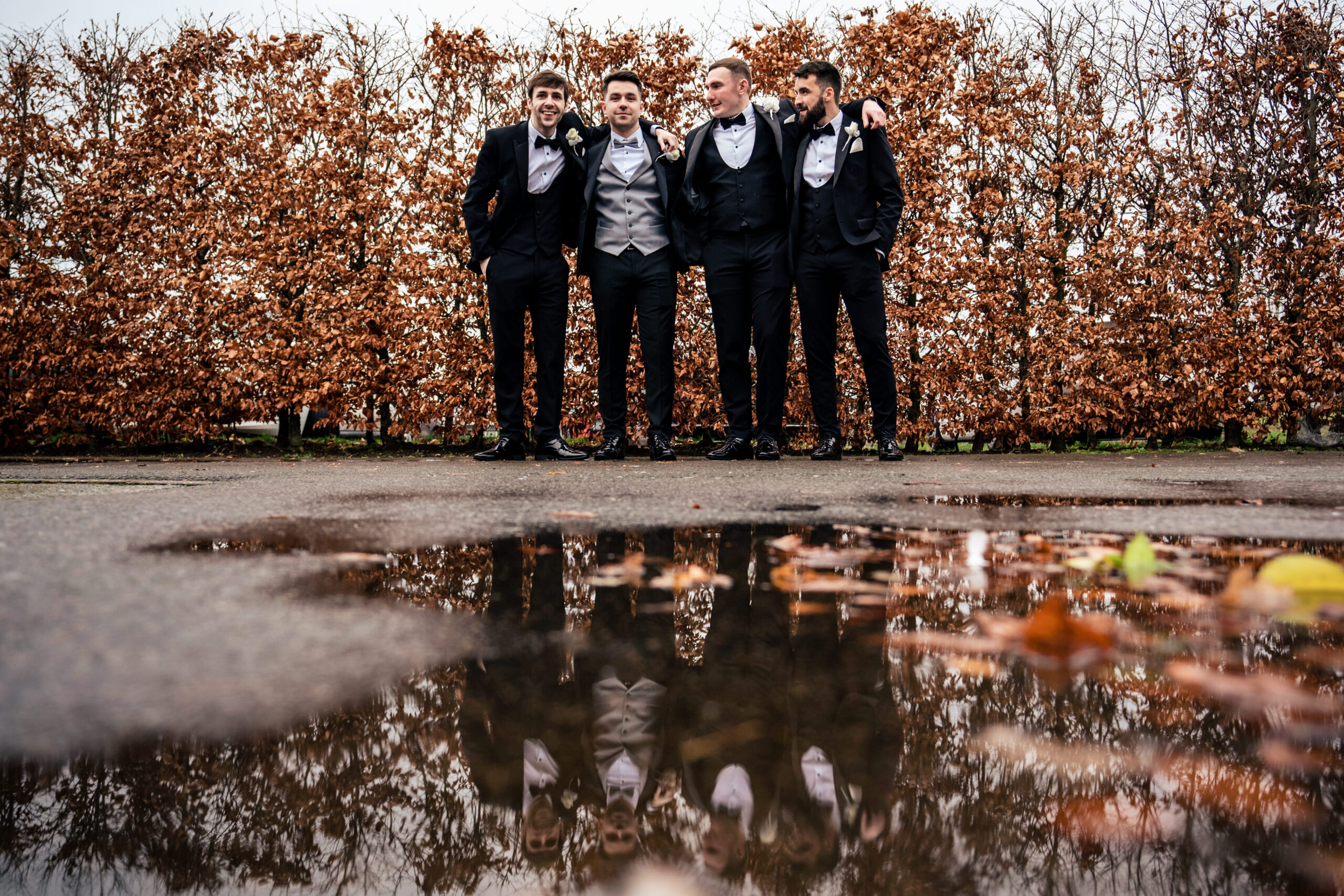 Jamie and his groomsmen in black tie reflected in a rain puddle in front of a copper leaved hedge at Merrydale Manor