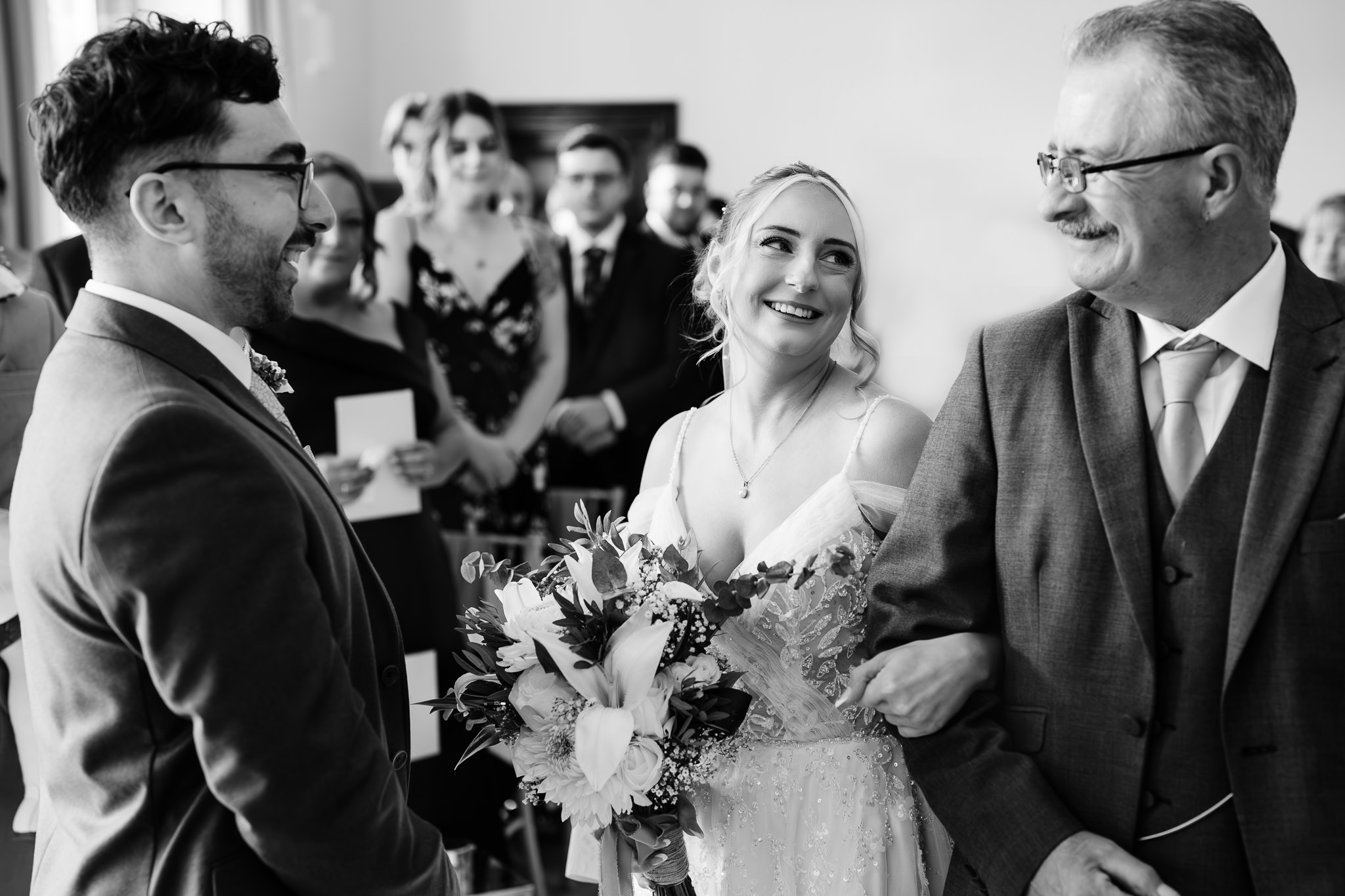 Black and white photo of the groom wiping away tears as the bride arrives on her dad’s arm