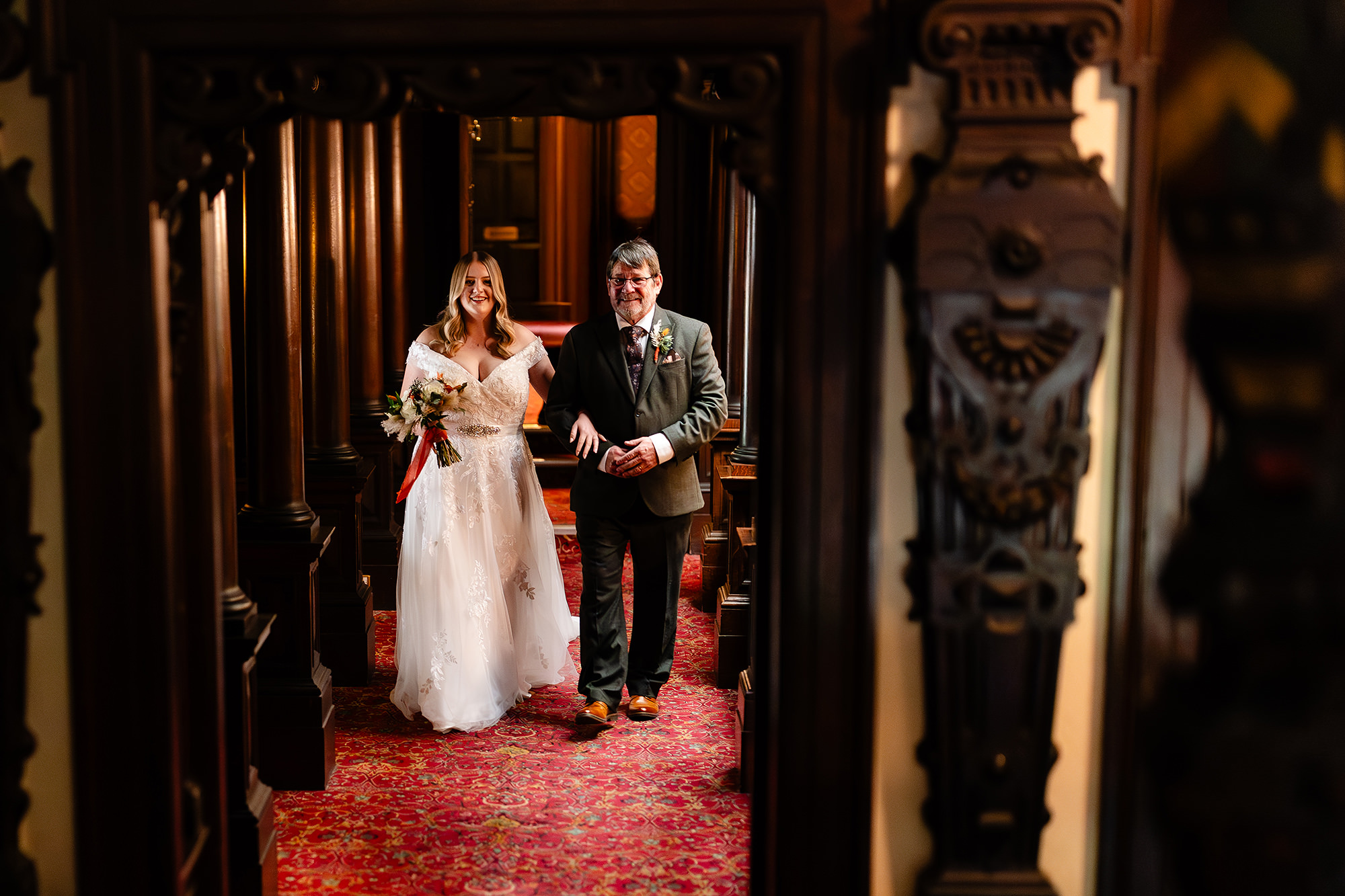 The bride and her dad walking arm in arm along Crewe Hall’s dark wood panelled corridor towards the ceremony room, Cheshire wedding photographer