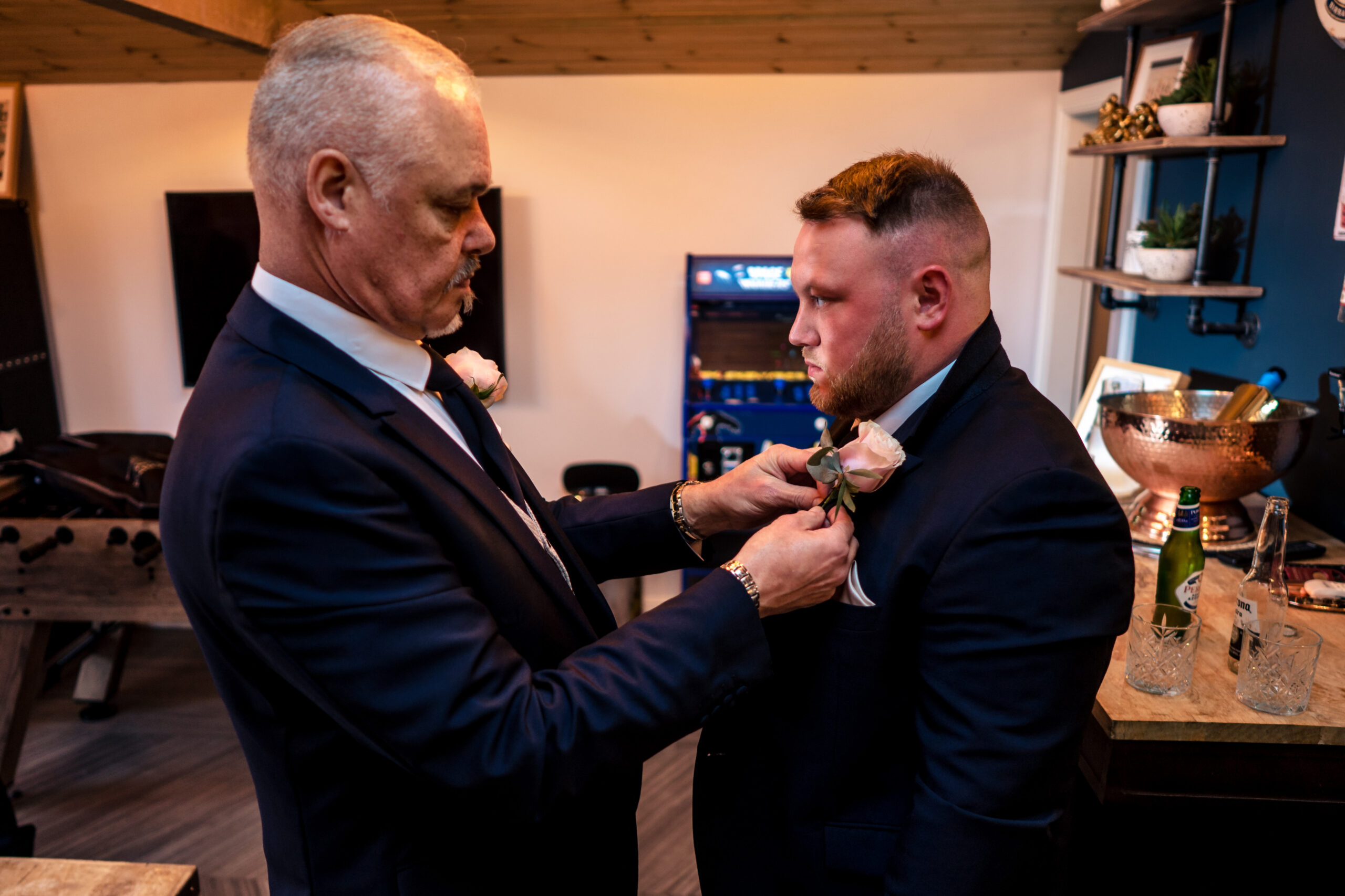 A pink rose buttonhole being pinned onto a groomsman's navy suit in the Man Cave