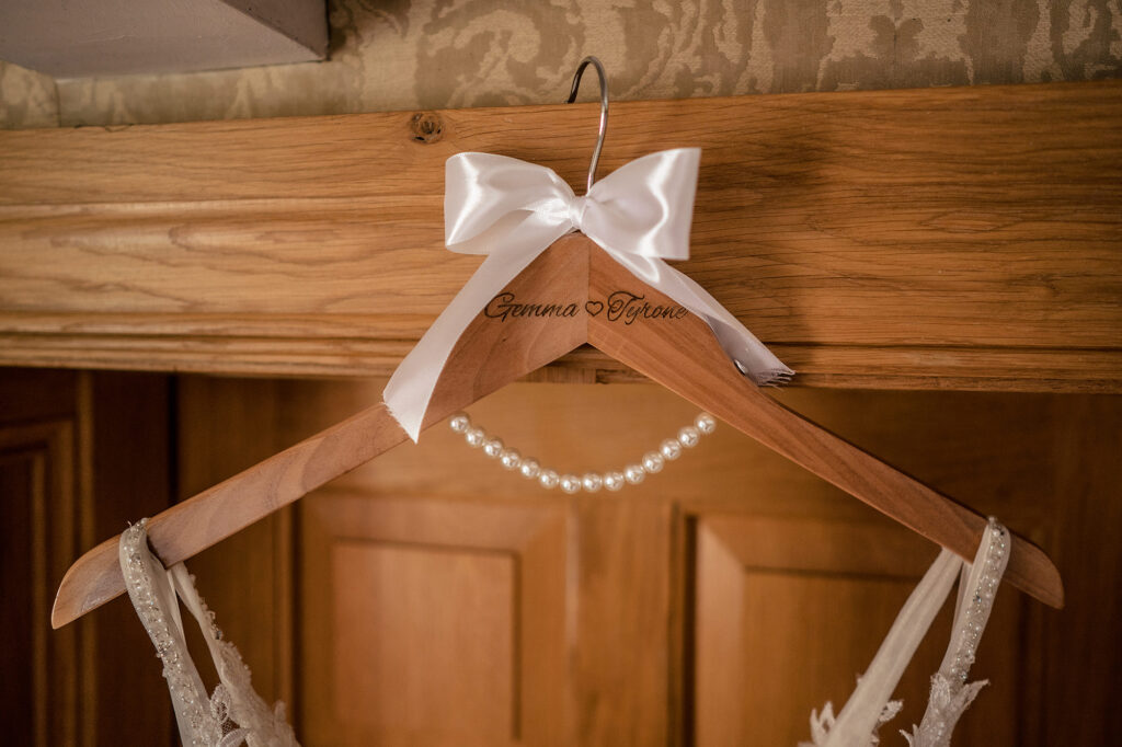 Wooden hanger engraved with Gemma and Tyrone’s names, tied with a white ribbon and a string of pearls, holding the wedding dress