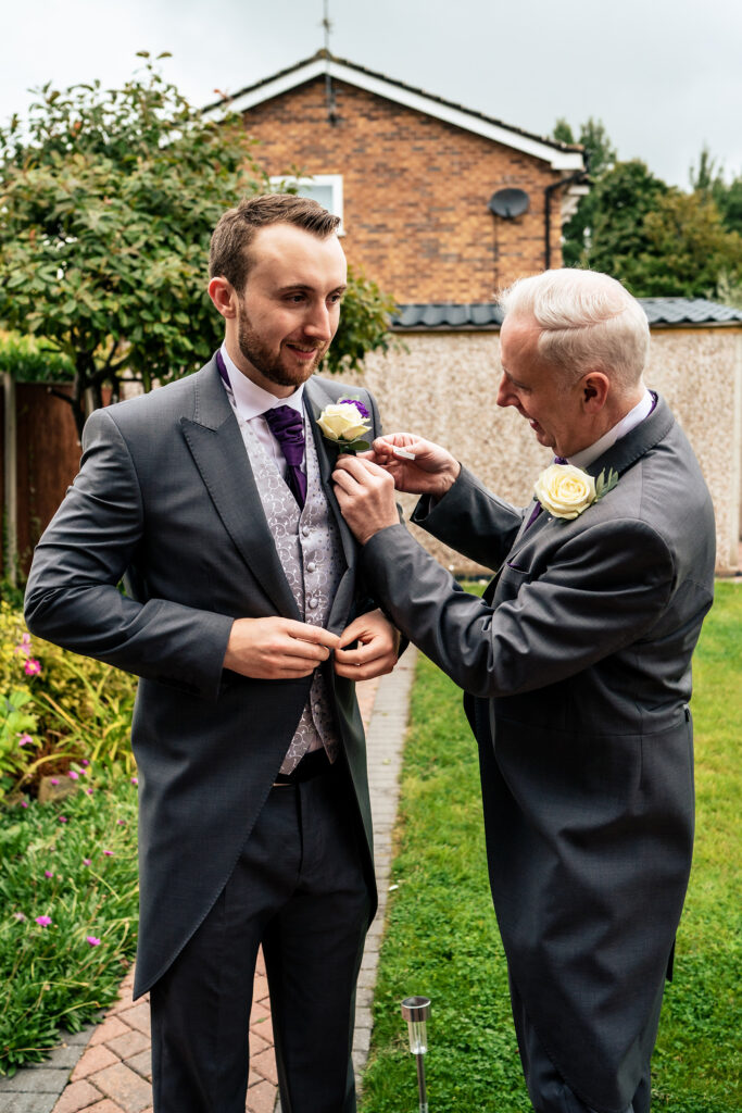 Groom's dad pinning a cream rose buttonhole onto his morning suit in the garden, Merrydale Manor wedding photographer