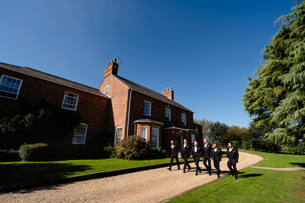 Groomsmen in black tie walking up the drive in front of the red brick main house under a blue sky, Swancar Farm wedding photographer