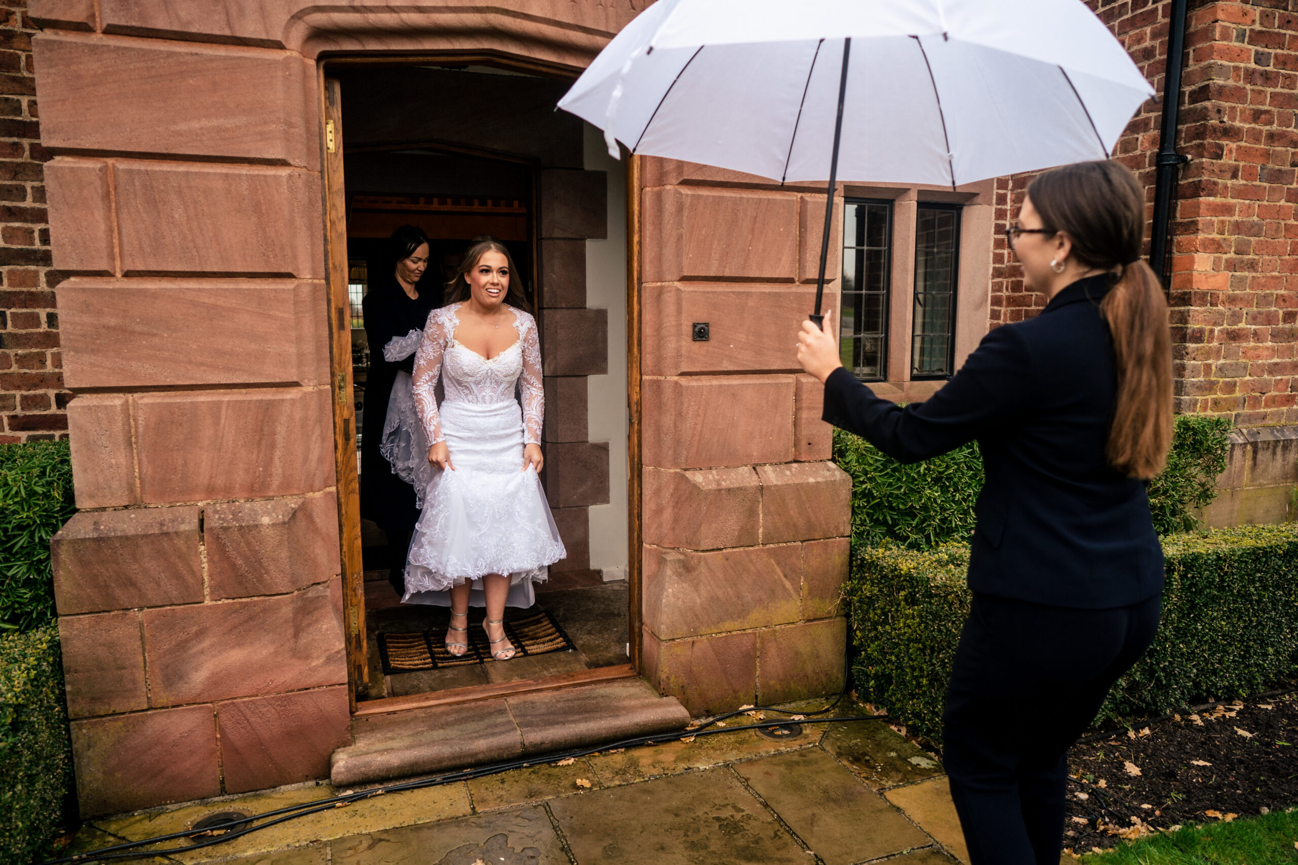 Abbie stepping out of a stone doorway under a white umbrella on a rainy December wedding day at Merrydale Manor