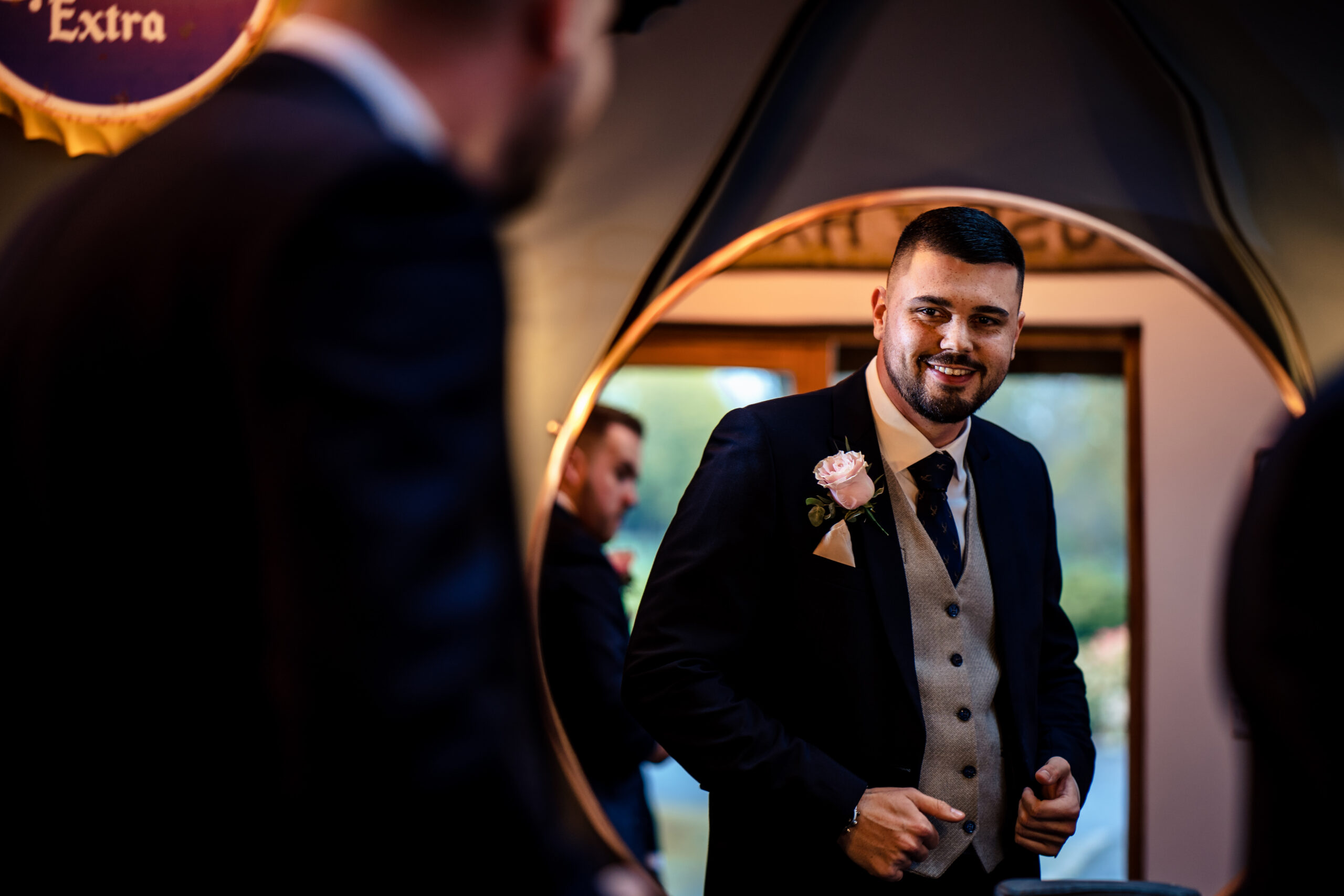 Dean in his navy suit and grey waistcoat reflected in a round mirror in the groom's room