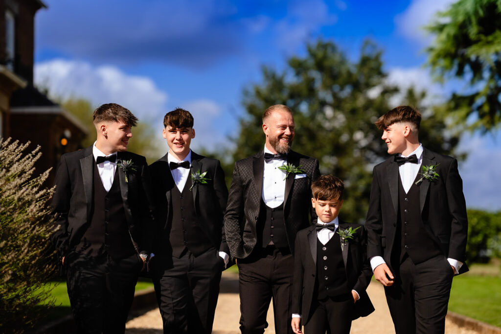 Glynn walking with his groomsmen and a young boy in black tie in bright sunshine, Swancar Farm wedding photographer