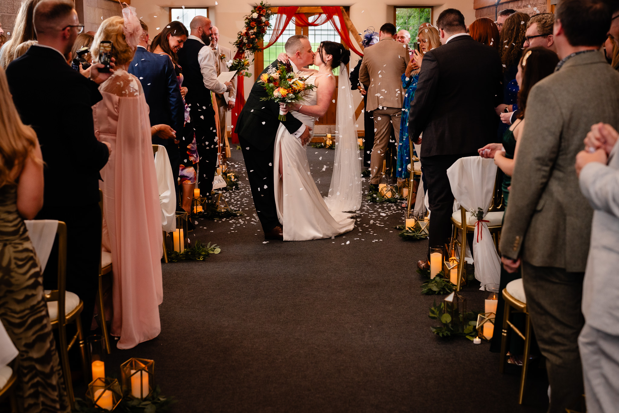 Bride and groom kissing halfway down the aisle as guests throw petals over them, Heaton House Farm wedding ceremony barn