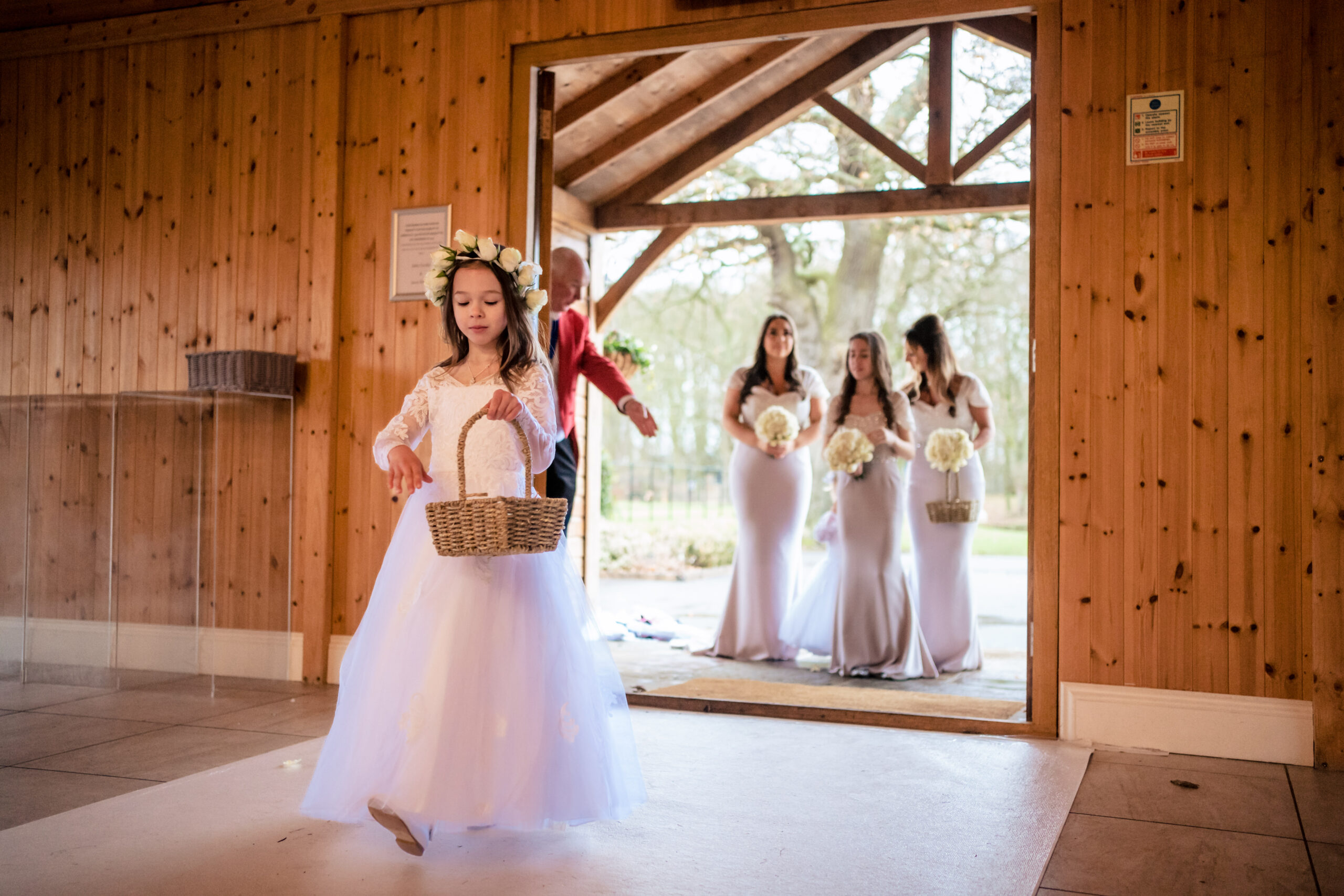 Flower girl in a flower crown carrying a wicker basket into the ceremony as the bridesmaids wait at the door, Merrydale Manor