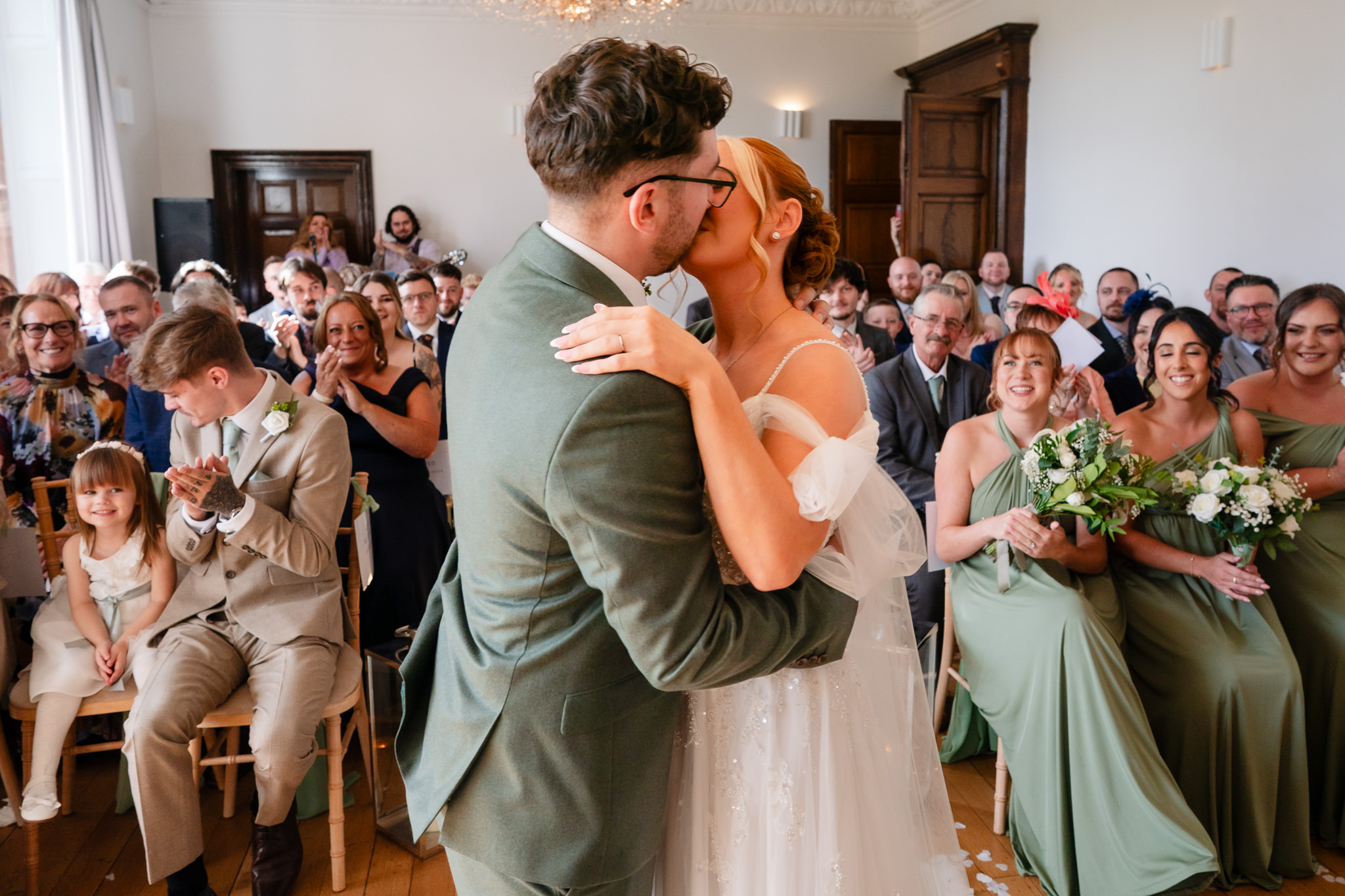 The couple’s first kiss as newlyweds, guests seated around them applauding at Salt House, Staffordshire wedding photographer