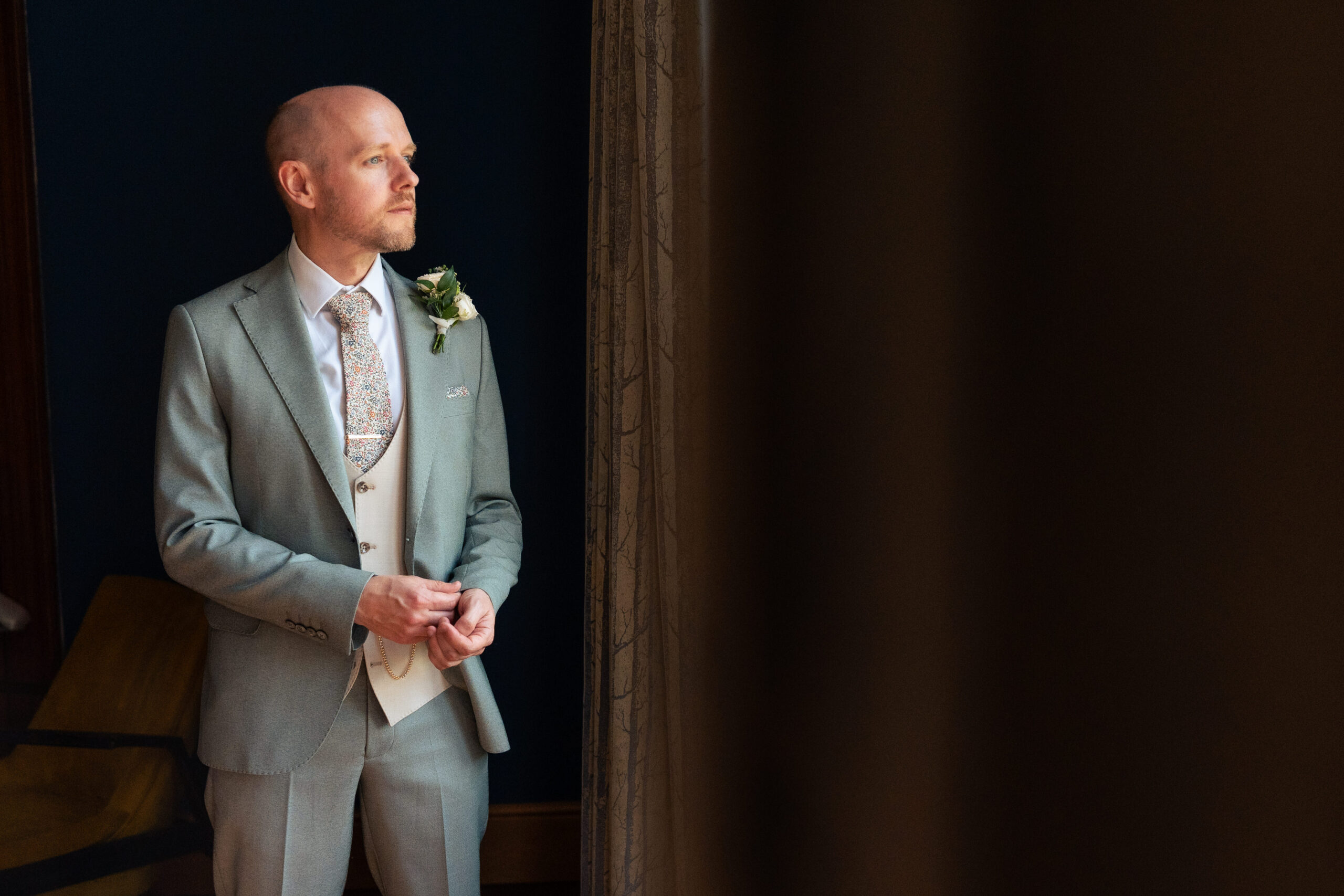 Groom in a sage green three piece suit standing by a window at Salt House ahead of his wedding