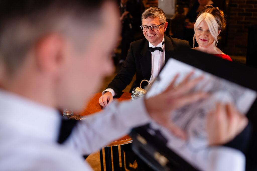 Caricature artist sketching two smiling guests in black tie before the ceremony, Swancar Farm Country House, Nottingham wedding photographer