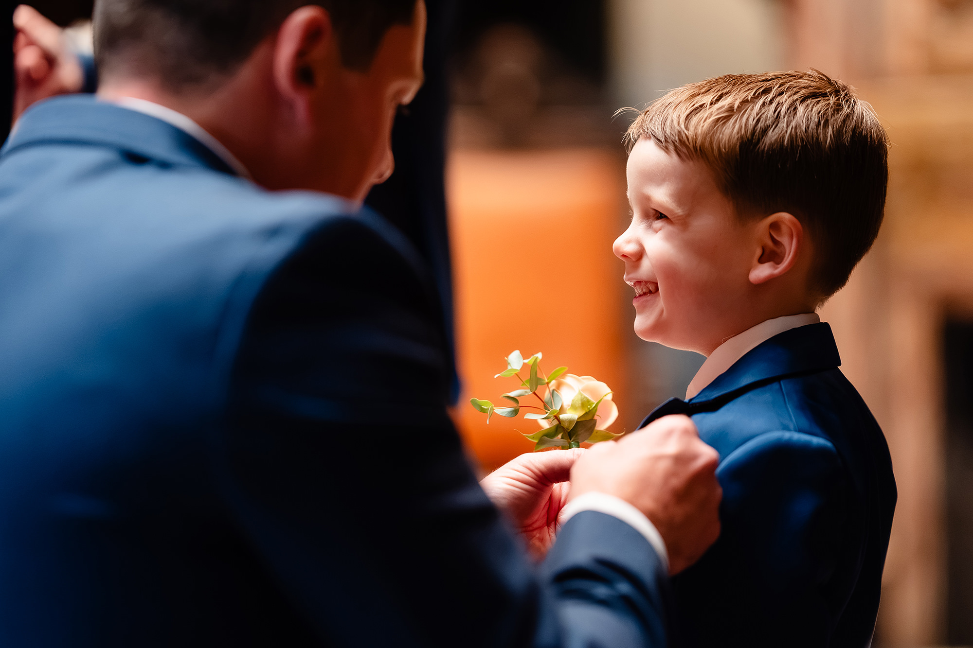A buttonhole being pinned onto a small boy’s navy suit while he grins up at the man doing it