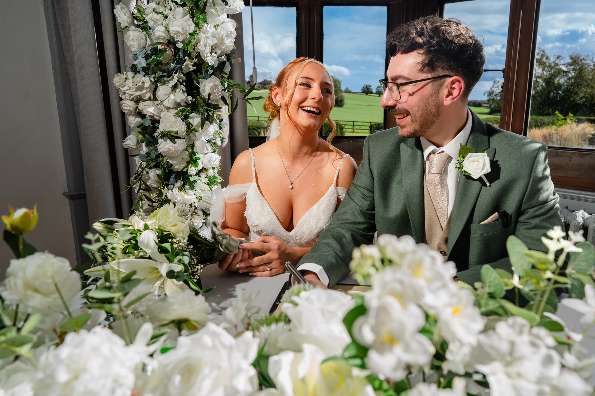 Bride and groom holding hands while signing the register, surrounded by white florals