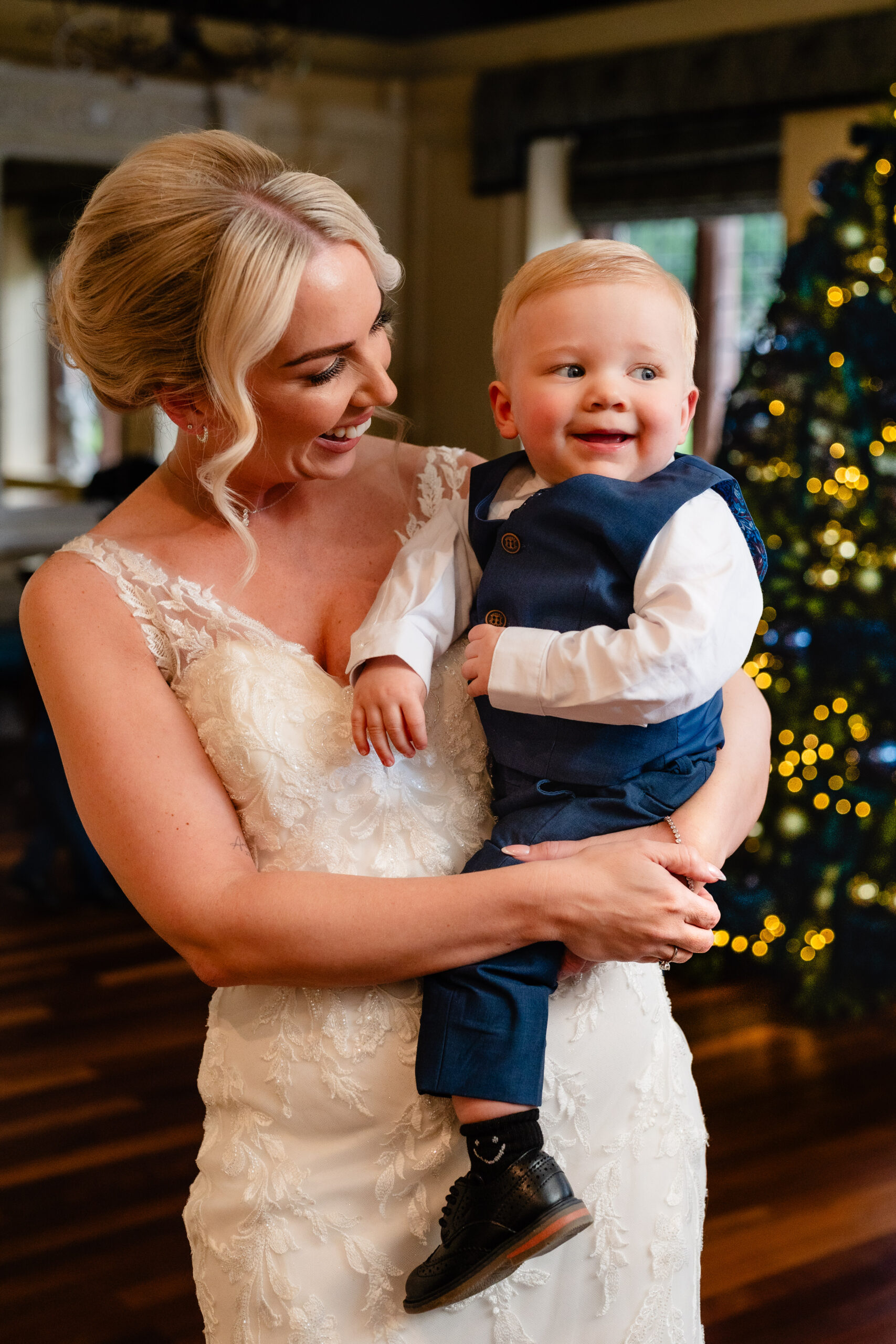 Bride in her wedding dress holding her baby son in a navy waistcoat in front of the Christmas tree, Colshaw Hall winter wedding