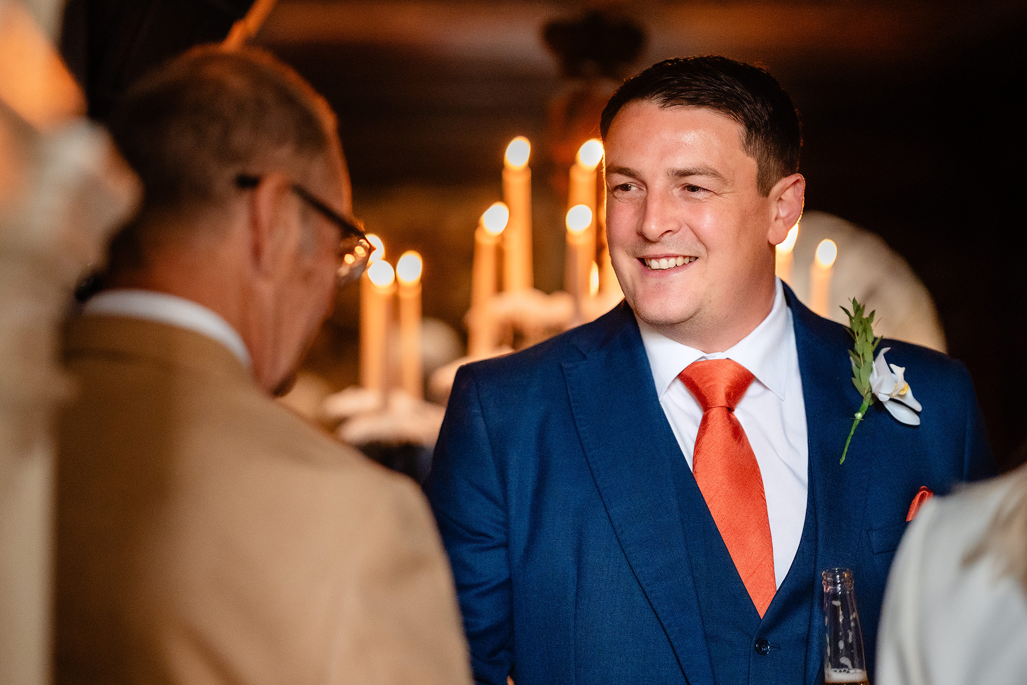 The groom in a navy three piece suit and orange tie laughing with a guest beside a bank of candles at Crewe Hall