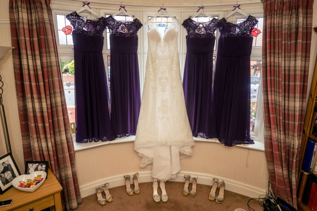 Wedding dress hanging in a bay window between four purple bridesmaid dresses with shoes lined up below, Merrydale Manor wedding photographer