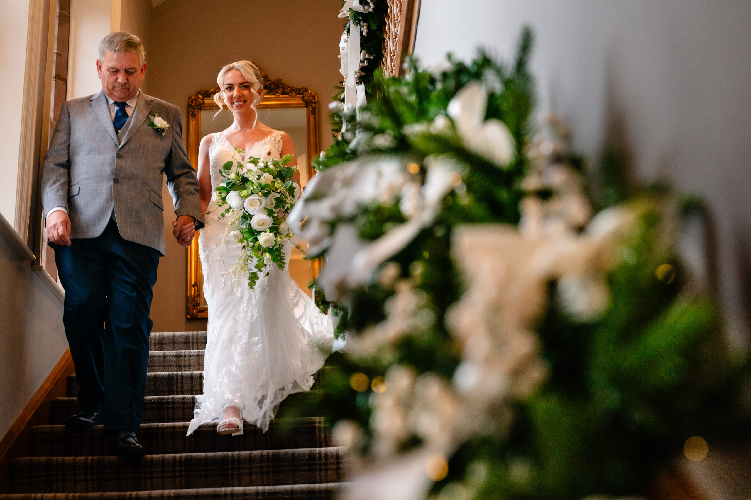 Bride walking down the staircase hand in hand with her dad past Christmas greenery at Colshaw Hall, Cheshire wedding photographer