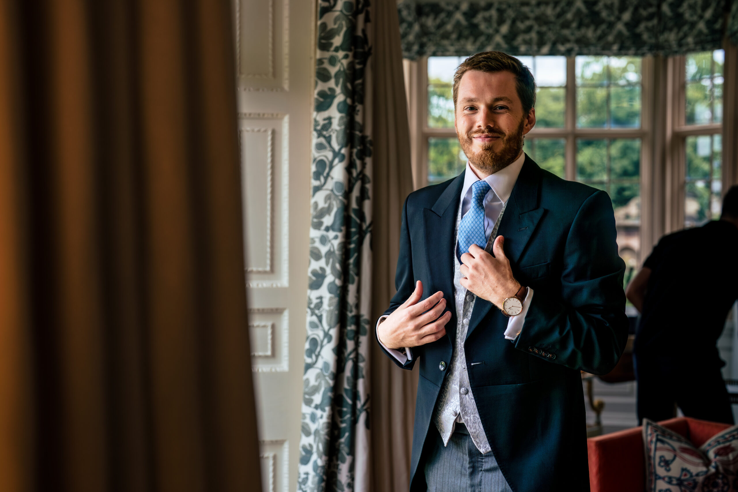 Groom straightening his tie by a tall window in the house at Dorfold Hall