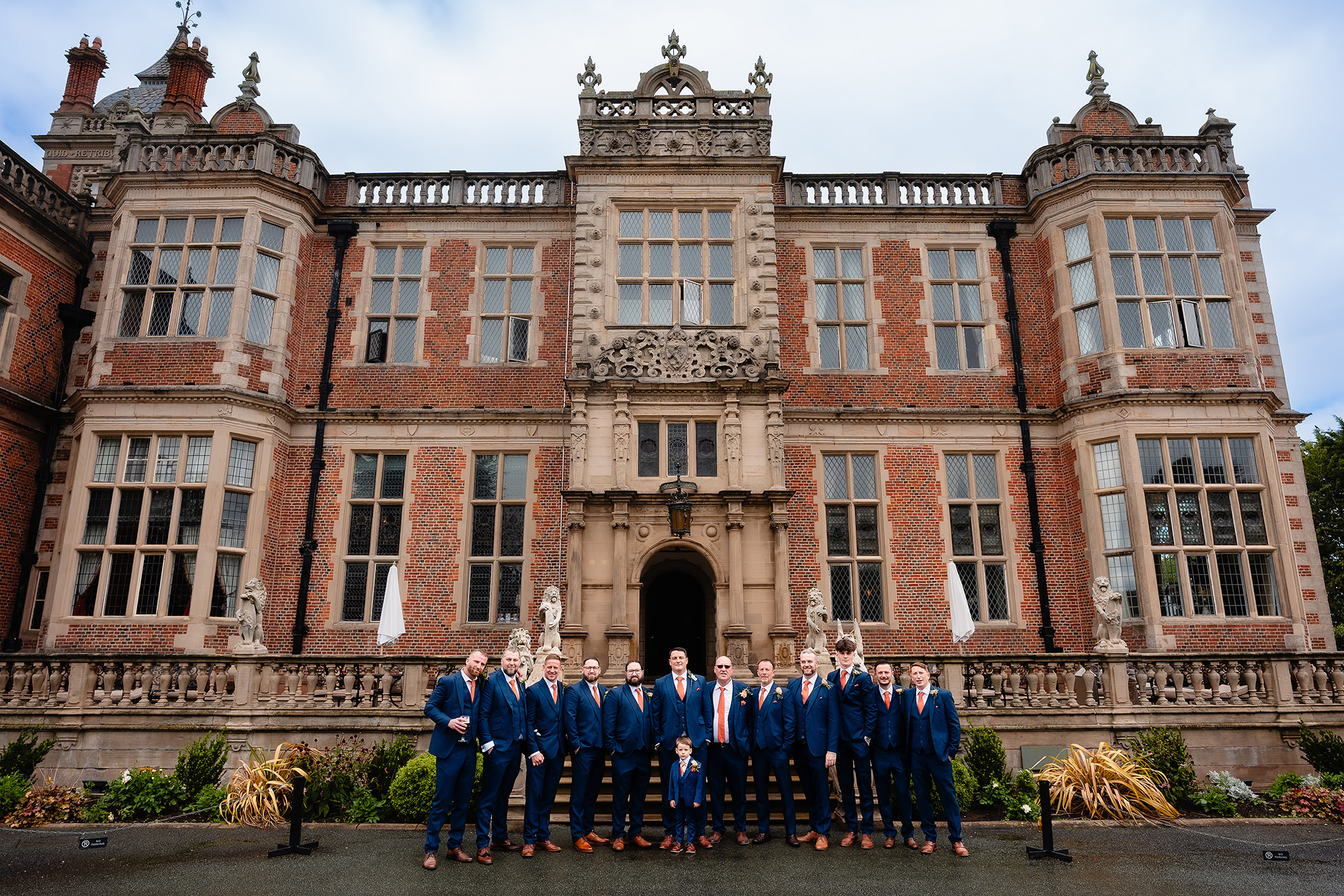 The groom and a long line of men in navy suits and orange ties lined up across the steps at Crewe Hall