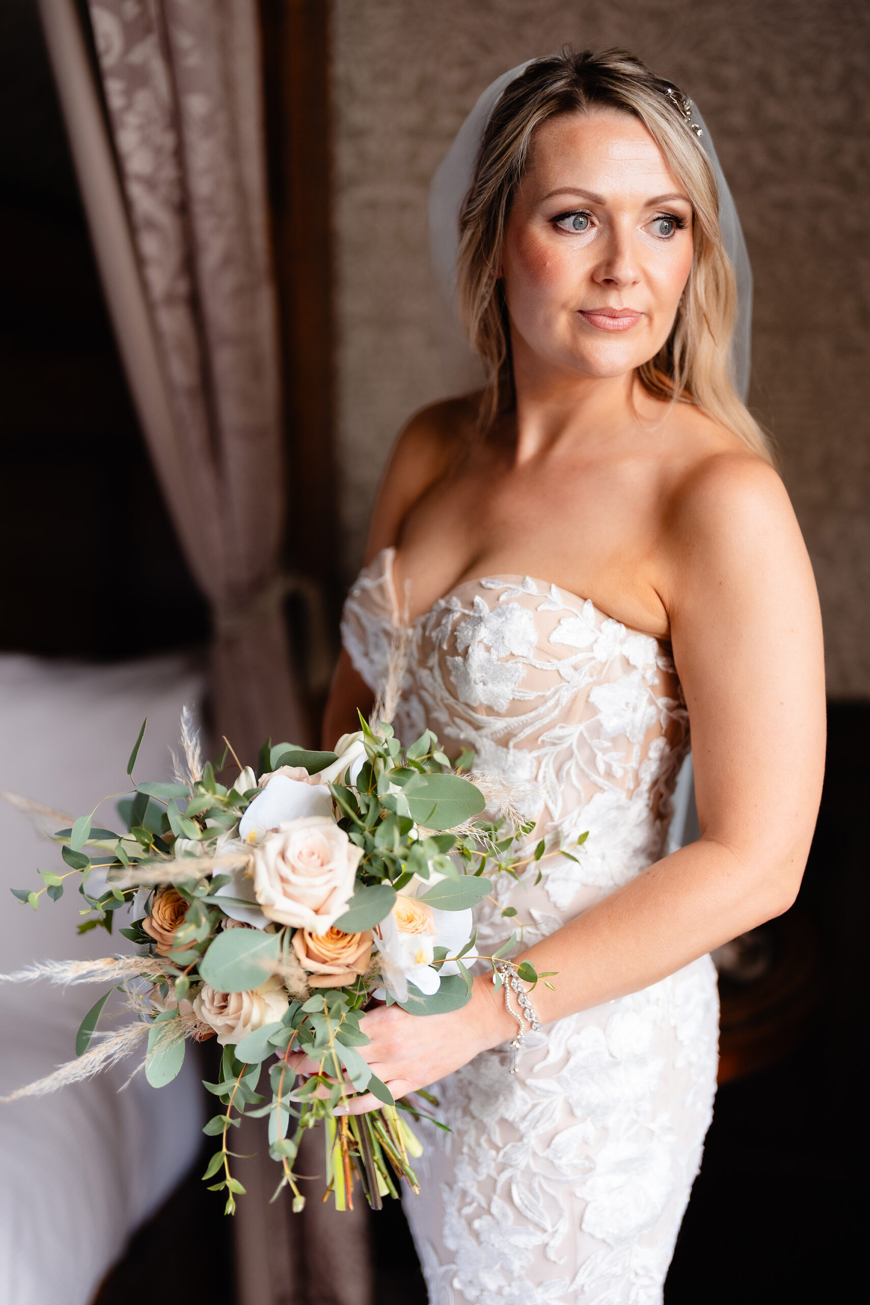 The bride in a strapless lace fit and flare dress holding a peach and eucalyptus bouquet by a window at Crewe Hall