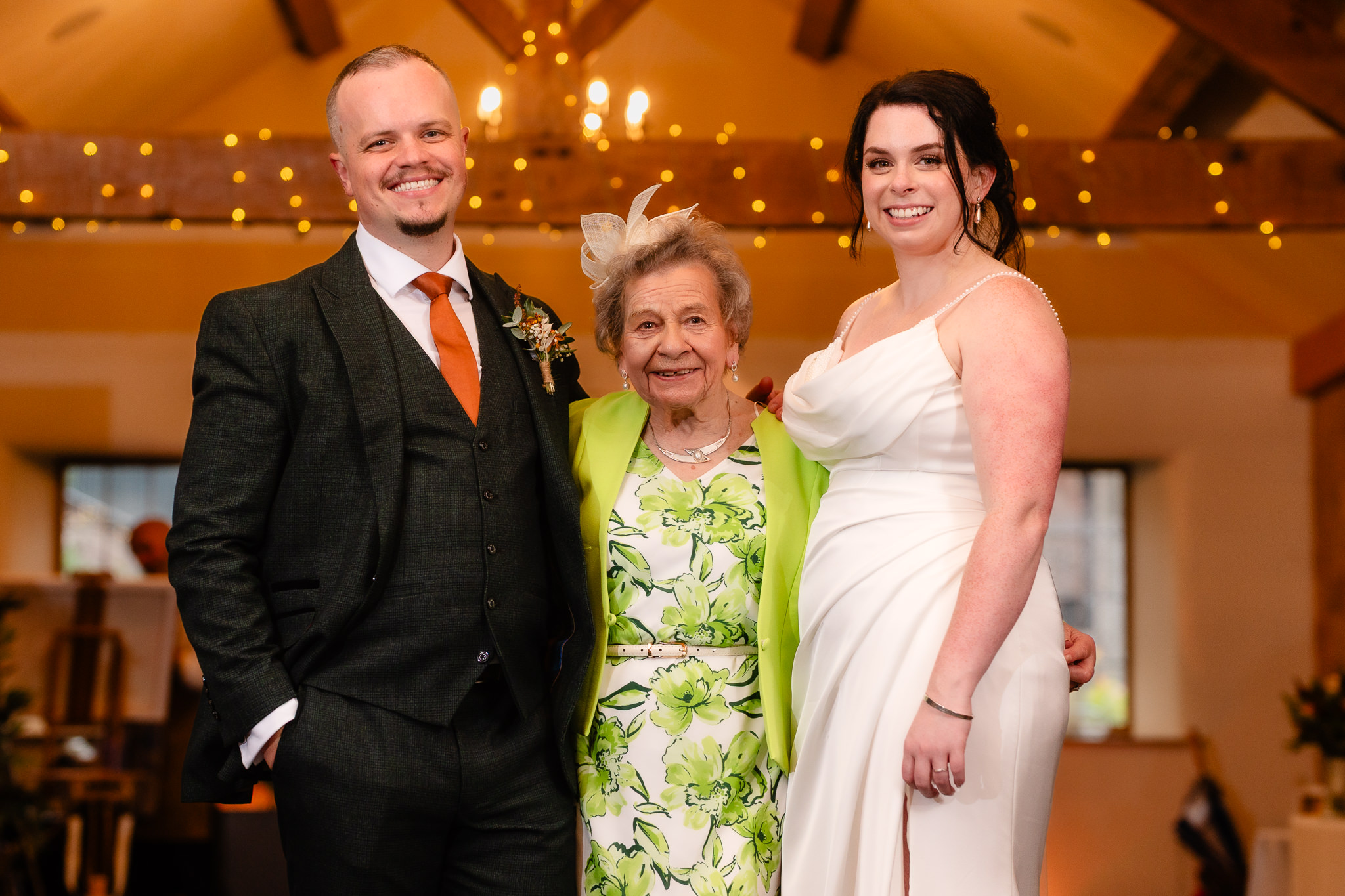 Bride and groom either side of the bride's grandmother in a green floral dress under the barn beams, Heaton House Farm wedding
