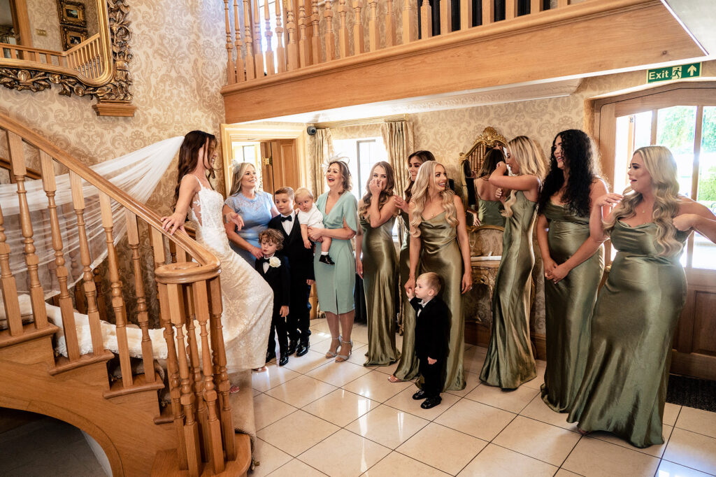 Bridesmaids in olive green satin and family reacting as Gemma comes down the staircase in her wedding dress at Merrydale Manor