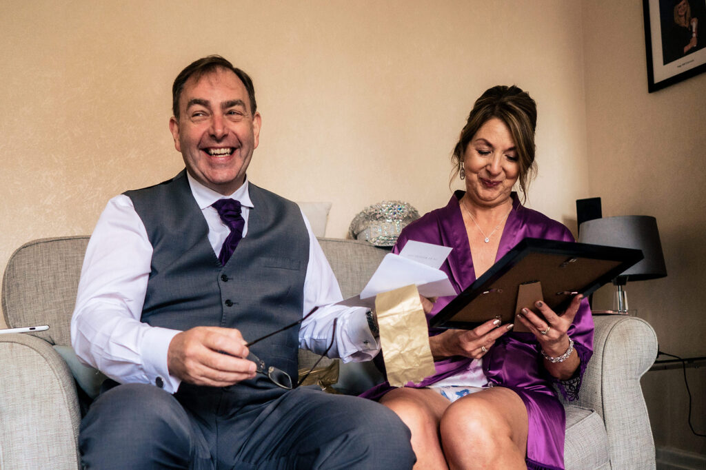 Bride's mum and dad laughing as they open a framed gift and a handwritten letter on the wedding morning, Cheshire wedding photographer