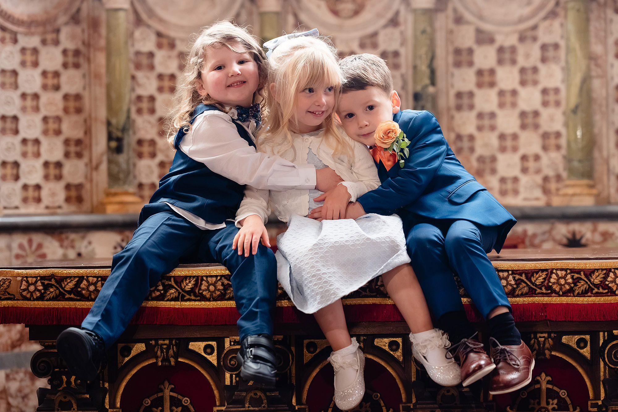 Three small children squashed together on an ornate bench, two boys in navy suits and a girl in white