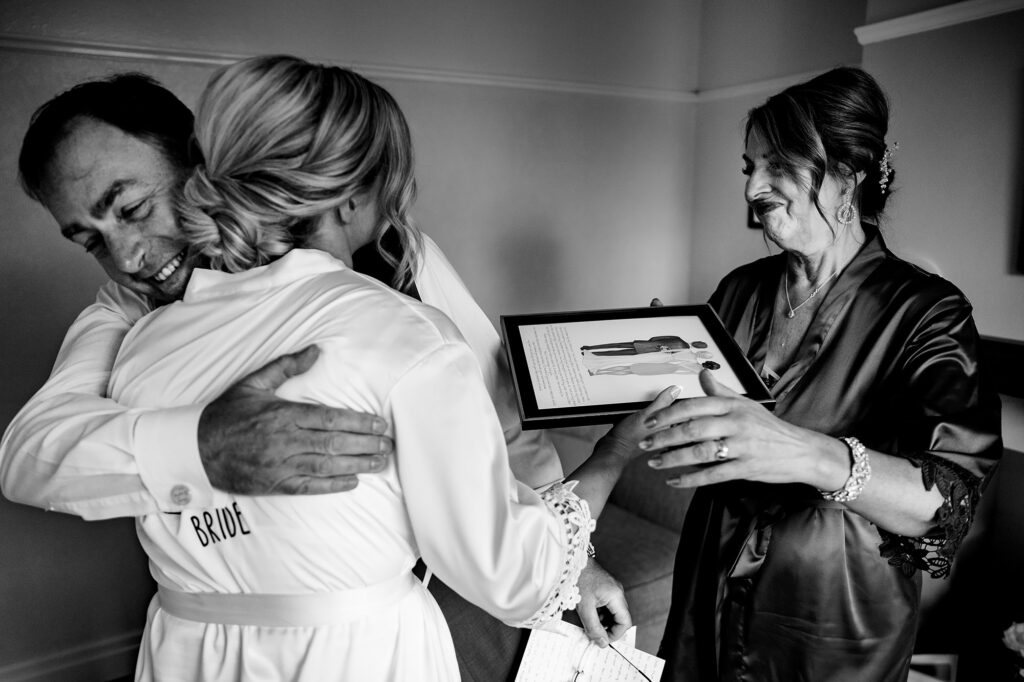 Bride in her robe hugging her dad while her mum holds a framed illustration, black and white, Merrydale Manor wedding photographer