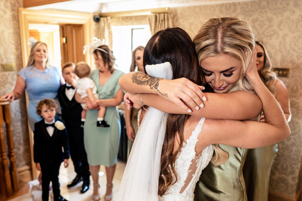 Gemma hugging a bridesmaid in the hallway after her dress reveal, with page boys and family watching