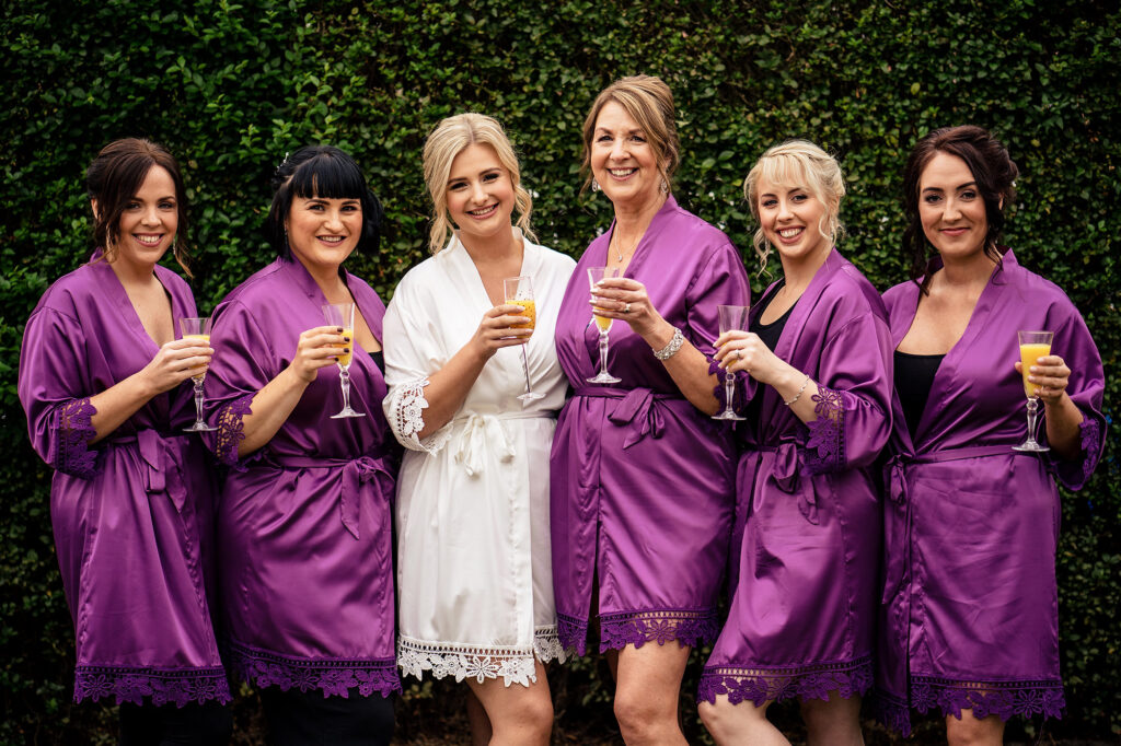 Bride in a white robe with her bridal party in purple satin robes raising drinks in the garden, Cheshire wedding photographer