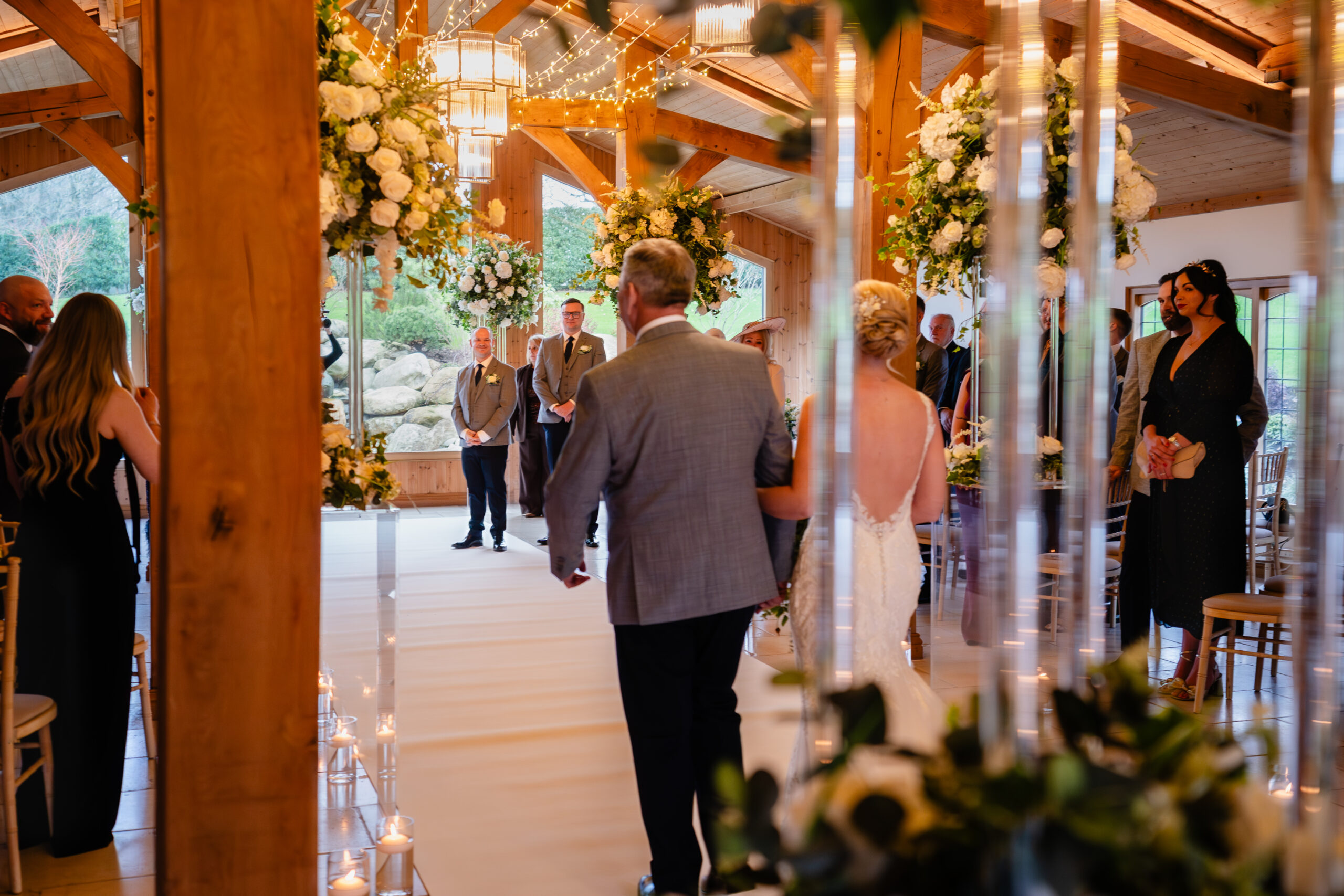 The bride heading down the white aisle runner between tall white and green floral pillars and candles, Colshaw Hall wedding photographer